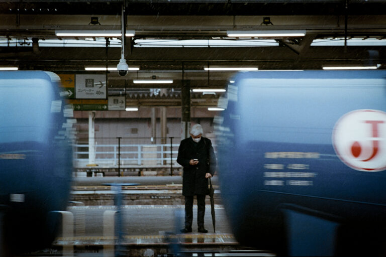 A lone person on a railway platform between two passing blue trains.