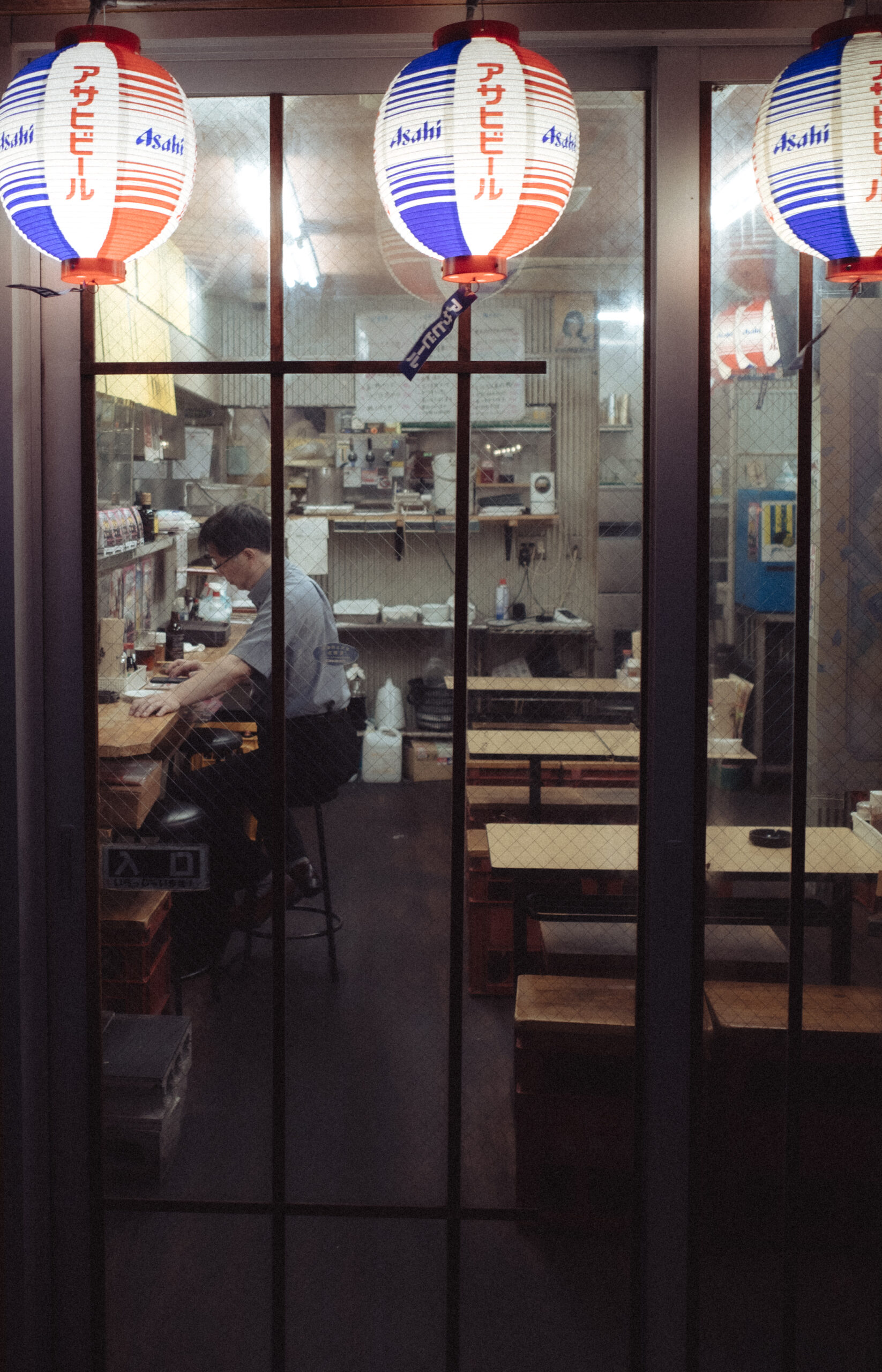 A man sitting alone at a counter beneath illuminated Japanese beer lanterns.