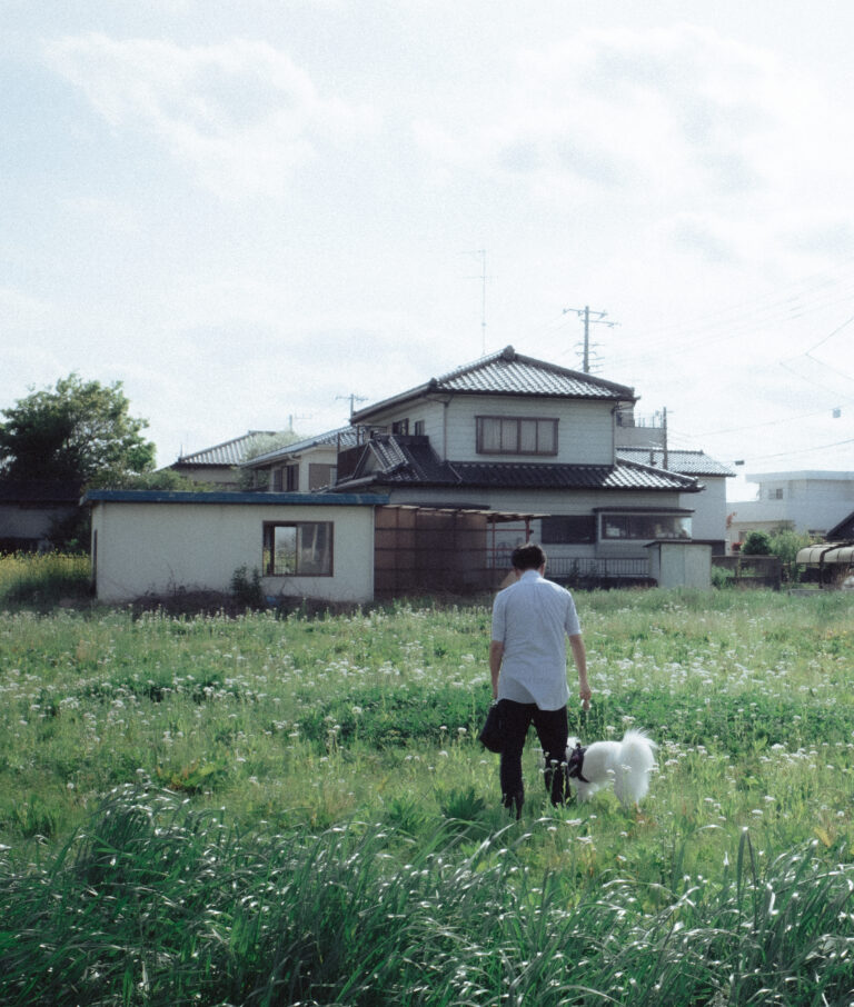 A person and a dog crossing a field toward a house.